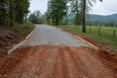 Long rural driveway with fresh concrete in the foreground giving way to a rutted red-clay gravel track toward a farmhouse